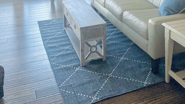 Sala de estar con un sofá de cuero blanco, un sillón blanco y un tapete azul estampado. Hay un ventilador de techo, una ventana con vistas a la playa y una puerta que da al exterior.