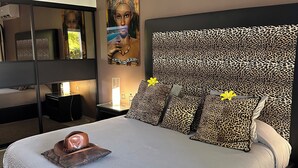 A bedroom with a large bed, a headboard with a leopard print pattern, a nightstand with a lamp, and a painting on the wall. - Studio Master   Residence Adam et  Eve (Baie Orientale)