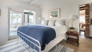 A bedroom with a bed, bedside lamps, a framed picture, and a view of the outdoors. - Steps to Beach + Historic Cottage | Primary Balcony + Hammock | Sandpiper (Hilton Head Island)