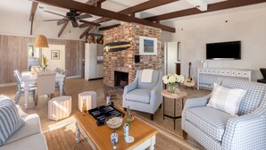 A spacious living room with a brick fireplace, a dining area, and a television mounted on the wall. - Steps to Beach + Historic Cottage | Primary Balcony + Hammock | Sandpiper (Hilton Head Island)