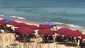 Una playa con sombrillas rojas y sillas de descanso, un muelle al fondo y gente nadando en el agua.