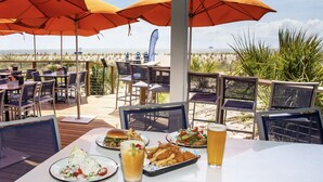 Ein Außenbereich zum Essen mit Tischen, Stühlen und Sonnenschirmen, mit Blick auf den Strand.