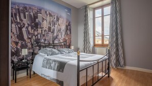 Un dormitorio con un mural de paisaje urbano, una cama con cubrecama blanco, una mesita de noche con lámpara y una ventana con cortinas estampadas.