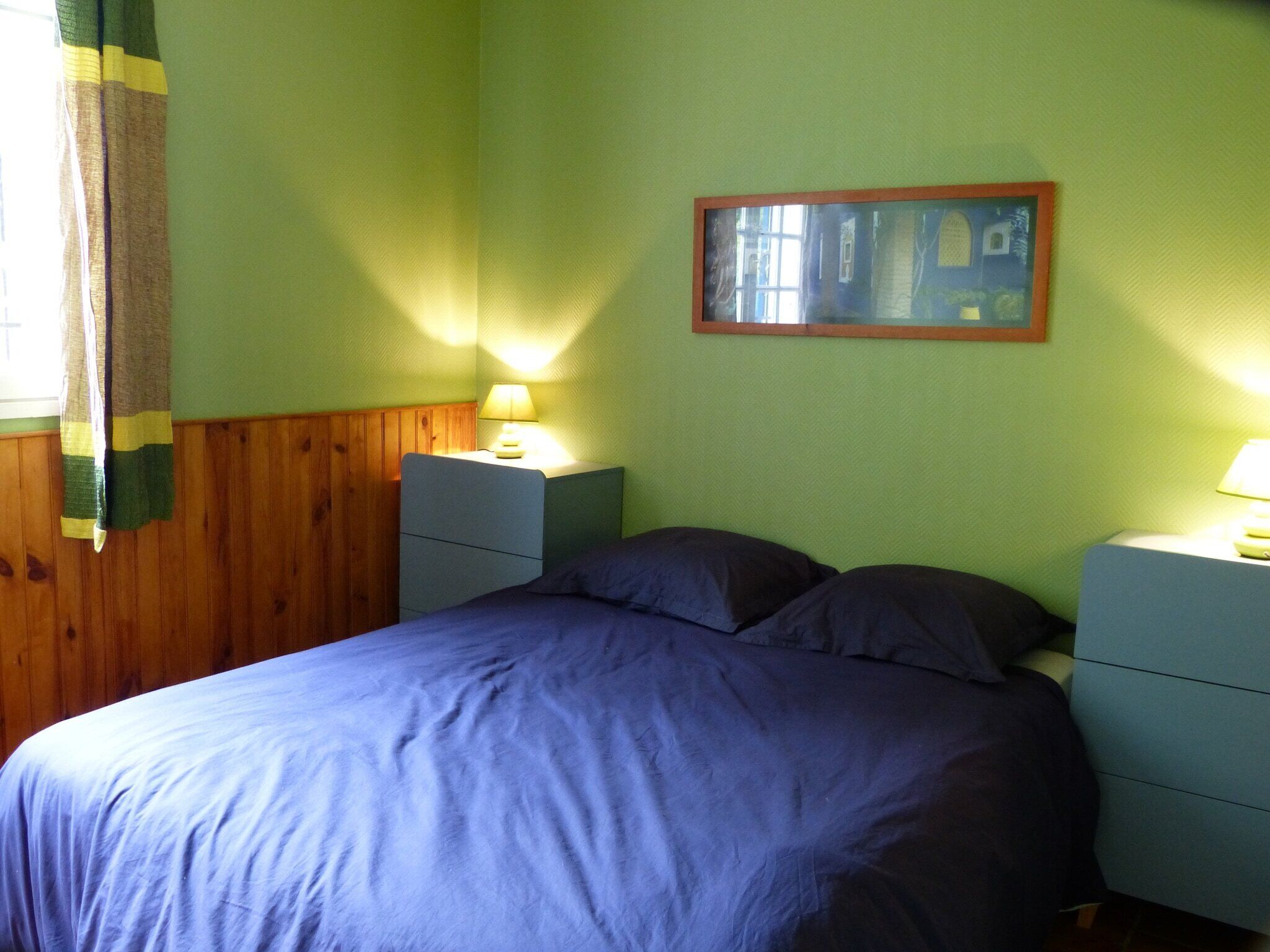 A bedroom with a bed, two bedside tables, a nightstand, a lamp, and a framed picture on the wall.