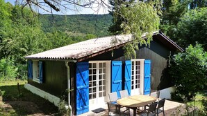 Une maison de campagne en bois, avec des volets bleus, une terrasse carrelée et une table entourée de chaises.