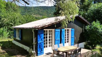 Une maison de campagne en bois, avec des volets bleus, une terrasse carrelée et une table entourée de chaises.