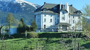 Un grand bâtiment blanc, doté de nombreuses fenêtres et d’un toit en pente, entouré de verdure et d’arbres, avec une montagne enneigée en arrière-plan.