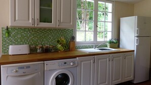 Une cuisine avec des meubles blancs, un lave-linge, un évier et une fenêtre donnant sur des arbres.