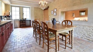 A dining area with a wooden table and chairs, a chandelier, and a kitchen in the background. - Elegant Normandy Retreat (Lantheuil)