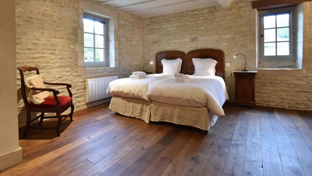 A bedroom with a stone wall, wooden floor, a bed with white linens, a wooden chair, and two windows. - Elegant Normandy Retreat (Lantheuil)