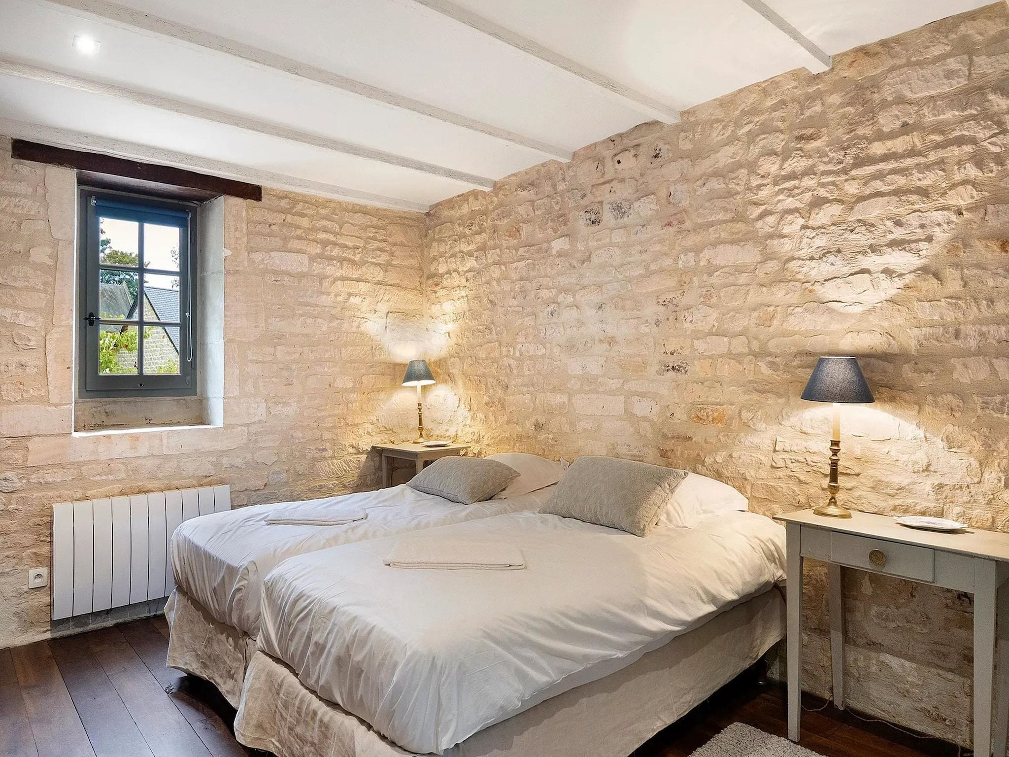 Une chambre avec un mur en pierre, un lit avec des oreillers, une table de chevet avec une lampe et une fenêtre donnant sur un paysage.