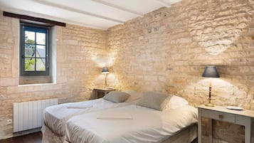 Une chambre avec un mur en pierre, un lit avec des oreillers, une table de chevet avec une lampe et une fenêtre donnant sur un paysage.