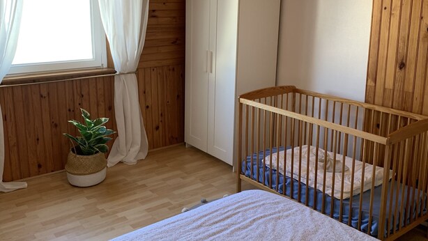 Een kamer met een babybedje, een kledingkast, een raam met gordijnen en een hanglamp.