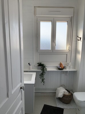 Een badkamer met een wit wastafelmeubel, een raam, een toilet en een klein plankje met toiletartikelen.