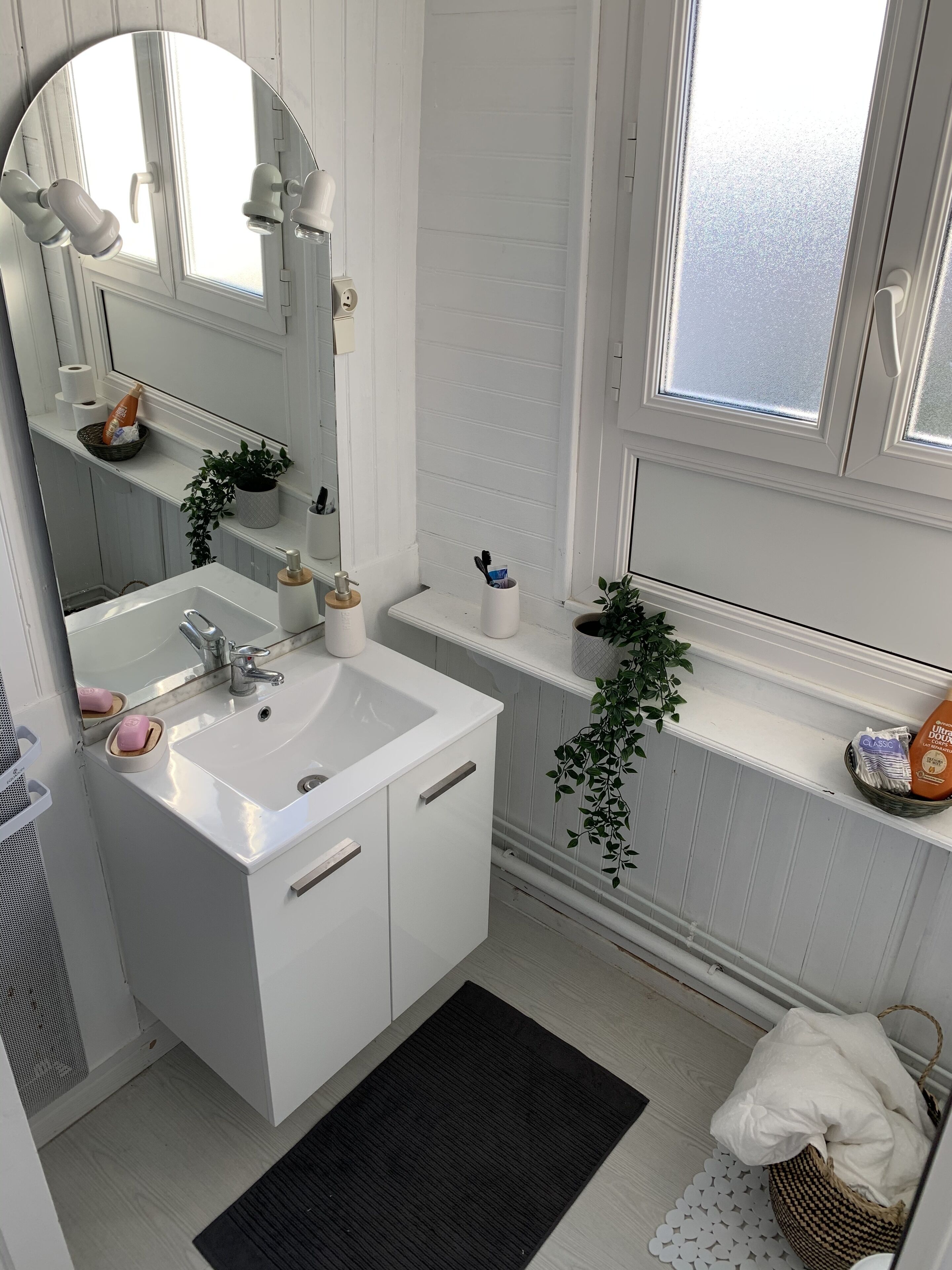 Een badkamer met een wit wastafelmeubel, spiegel en een venster met geëtste glas, plus een klein plankje met toiletartikelen.