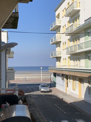 Een smalle straat met een geparkeerde auto, gebouwen met balkons en een helder uitzicht op het strand.