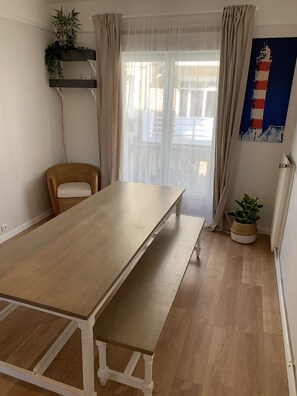 Een eetgedeelte met een lange houten tafel, een rieten stoel, een potplant en een schilderij van een vuurtoren aan de muur.