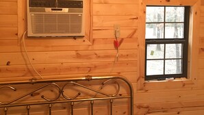 Interior de una cabaña de madera con una cama, una ventana, un aire acondicionado de pared y un cuadro con un paisaje de árboles.