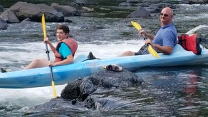 Dos personas haciendo kayak en un río con rocas y árboles al fondo.