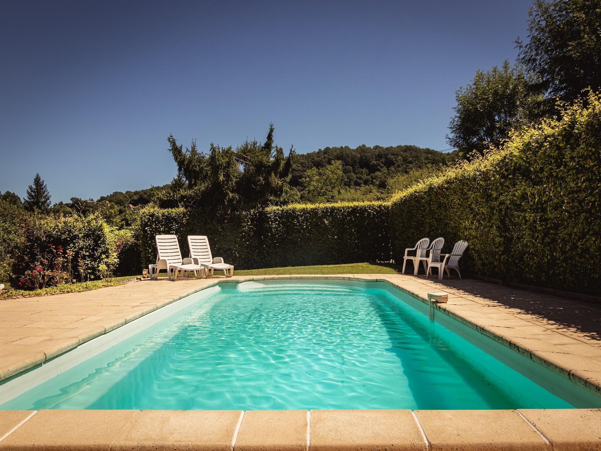 Una piscina rectangular con agua cristalina, rodeada por una terraza empedrada. Hay tumbonas a lo largo del borde de la piscina.