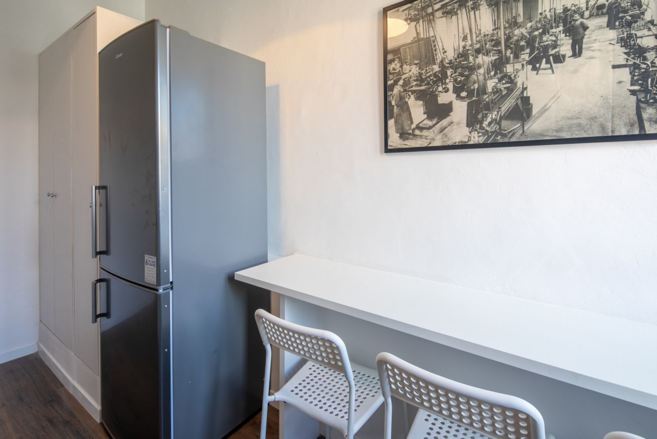 Een kleine keuken met een roestvrijstalen koelkast, een wit aanrecht en twee witte stoelen.