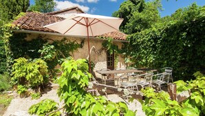 Un bâtiment rustique en pierre, doté d’un toit en tuiles, d’un patio couvert avec des chaises en métal et d’une végétation luxuriante.
