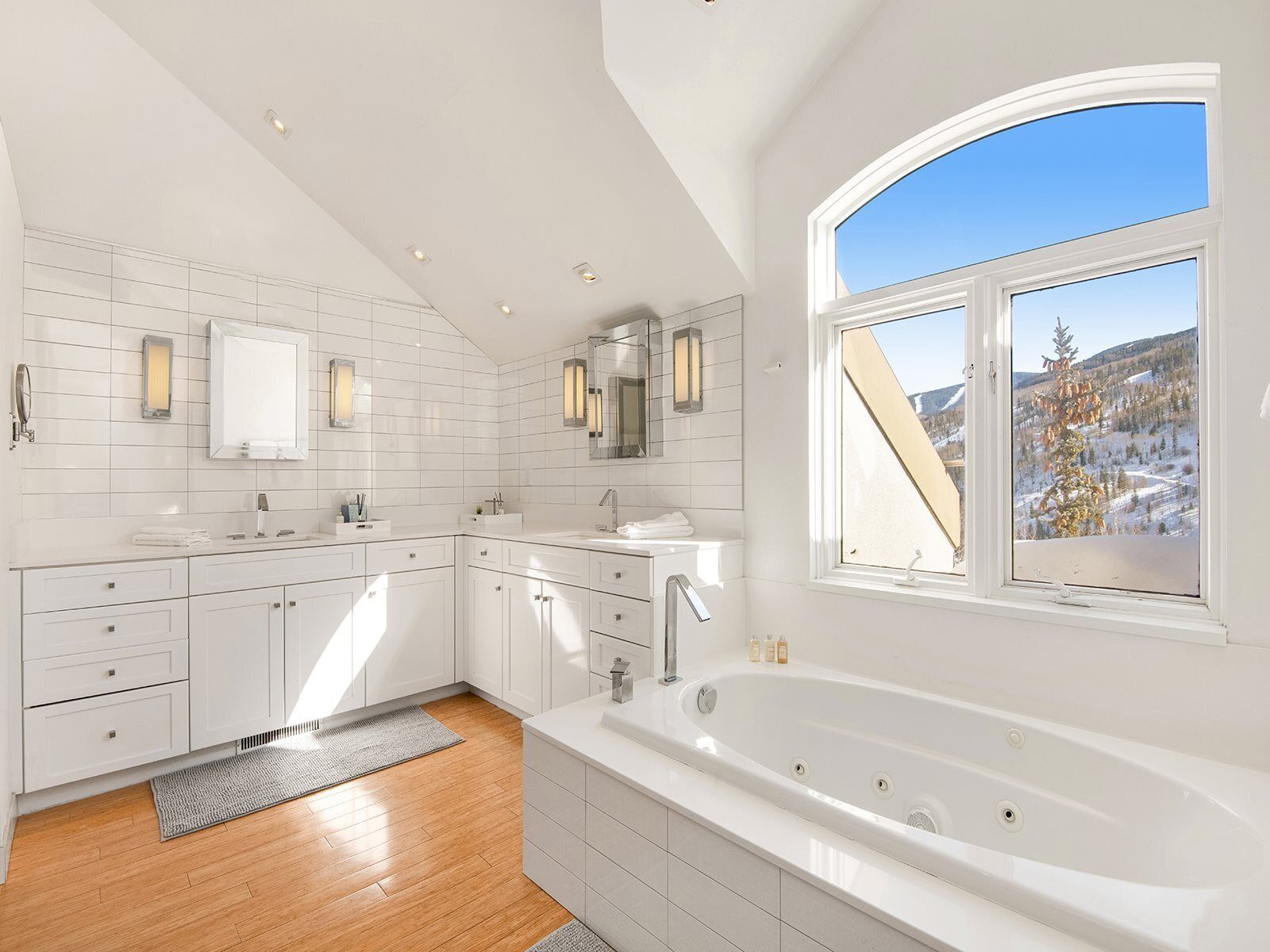 A modern bathroom with white tiles, a large bathtub, and a window with a view of a snowy landscape.