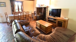 A cozy living room with a leather sofa, wooden coffee table, and a television. There is a brick fireplace and a dining area with wooden chairs and a table.