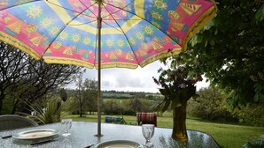 A table set outdoors with a colorful umbrella, a vase with flowers, and a view of a green landscape.
