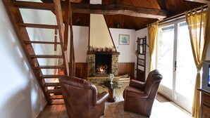 A cozy living room with a stone fireplace, wooden ceiling beams, a glass table, leather armchairs, and a television.