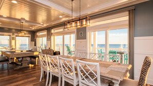 Un comedor espacioso con una mesa de madera y sillas blancas, ventanas amplias con vista al mar, e iluminación empotrada en el techo.