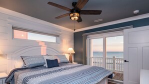 Un dormitorio con cama, ventilador de techo, mesita de noche, ventana y puerta que da a un balcón con vistas al océano.