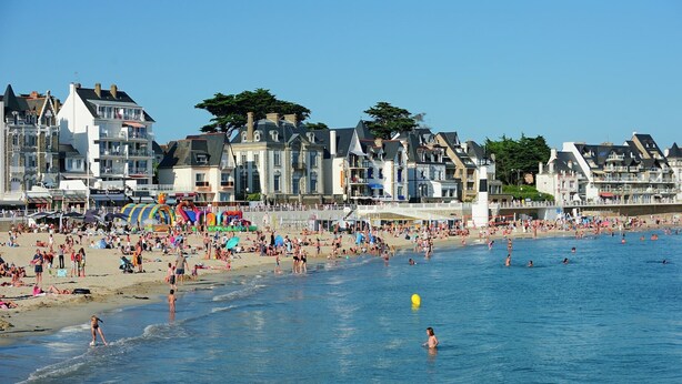 A beach with people swimming and sunbathing, and buildings in the background.