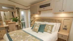 Une chambre avec un grand lit, un bureau et une chaise. Il y a une fenêtre avec des rideaux, une plante et une vue sur l’extérieur.