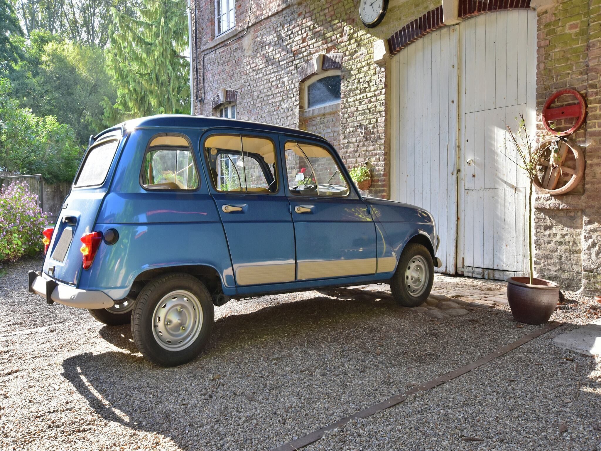 A vintage blue car parked in front of a rustic brick building with a white door and a clock on the wall.