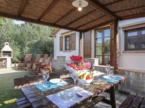 An outdoor dining area with wooden tables and chairs, a grill, and a view of a house with large windows and a garden. - La Villetta by Interhome (Riparbella)