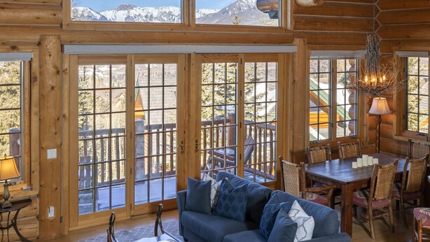 Un interior espacioso de cabaña de troncos con un amplio comedor, una sala de estar acogedora con sofá y mesa de centro, y grandes ventanales que ofrecen vistas a las montañas.