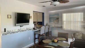 A dining area with a table set for four, a ceiling fan, a mounted TV, and a kitchen area in the background.