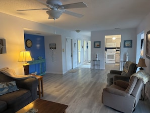 A living room with a ceiling fan, a clock, and a wall clock. There is a kitchen in the background with cabinets and a microwave. - Waterfront single home rental with private lanai, heated pool, and hot tub! (Venice)