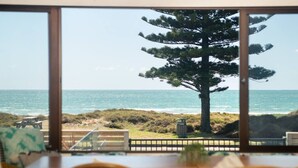 Vista da una stanza con ampie finestre su una spiaggia, un albero alto e un terrazzo in legno con mobili da esterno.