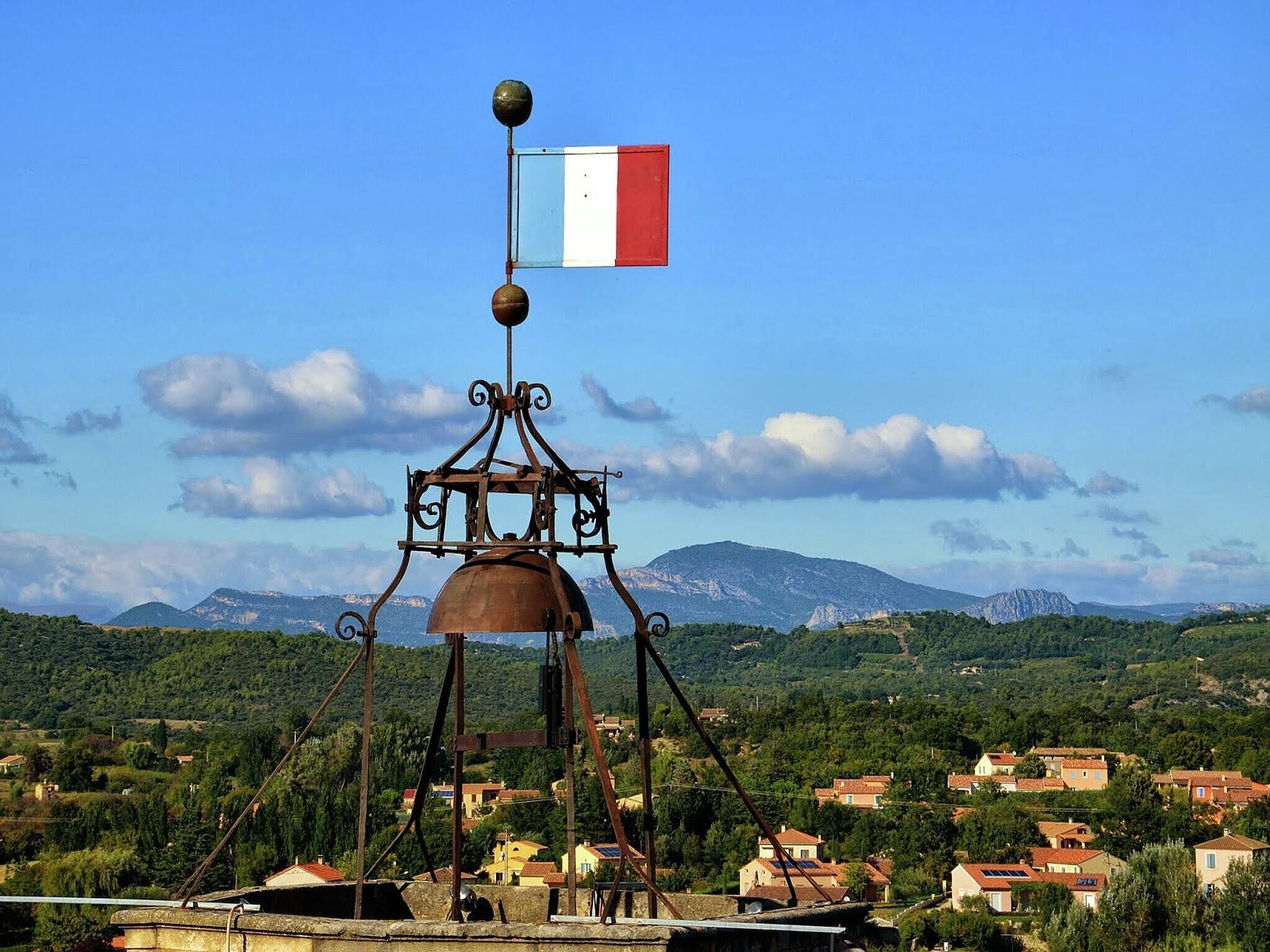 Un molino de viento con una bandera francesa, rodeado de un paisaje rural con casas y montañas.