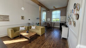 A spacious living room with a wooden floor, a sofa set, a dining table with chairs, and a kitchen area with a refrigerator and wall-mounted plates. - Holiday Home in Limousin near Forest (La Veytisou)