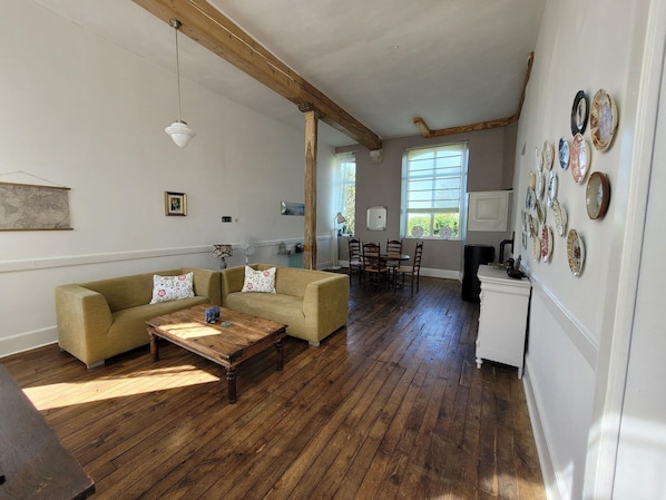 A spacious living room with a wooden floor, a sofa set, a dining table with chairs, and a kitchen area with a refrigerator and wall-mounted plates.