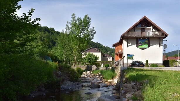 A riverside building with a balcony, surrounded by greenery and a small stream.