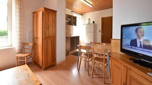 A room with a wooden ceiling, a dining table with chairs, a television, and a kitchen area with cabinets and a refrigerator.
