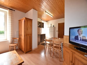 A room with a wooden ceiling, a dining table with chairs, a television, and a kitchen area with cabinets and a refrigerator.