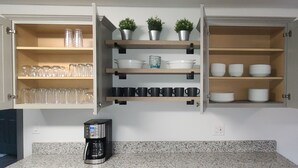 Una cocina con armarios abiertos que contienen cristalería, tazas y vajilla. Hay una cafetera sobre la encimera.