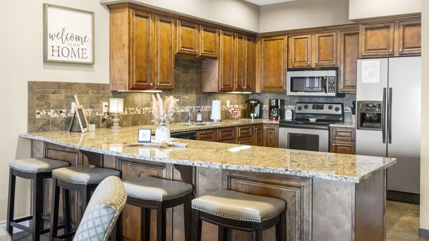 A kitchen with wooden cabinets, granite countertops, a central island with bar stools, a microwave, a refrigerator, and a coffee maker.