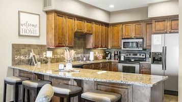 A kitchen with wooden cabinets, granite countertops, a central island with bar stools, a microwave, a refrigerator, and a coffee maker.