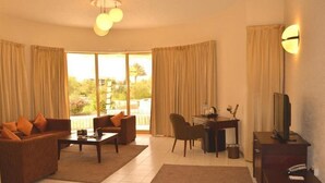 A living room with a sofa, coffee table, and TV stand, a desk with a chair, and large windows with curtains. - One to One Ain Al Fayda Resort (Al Ain)